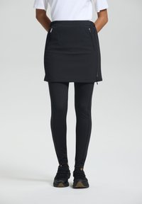 Musta skort, jossa on teksturoitu kuosi, varustettuna vetoketjullisilla taskuilla, yhdistettynä mustiin legginsseihin. Kengät ovat mustat ruskealla pohjalla.