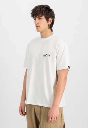 Joven con cabello oscuro despeinado lleva una camiseta blanca de Alpha Industries y pantalones cargo beige, de pie frente a un fondo claro y liso.