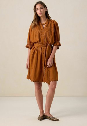Orangefarbenes Kleid aus verbrannter Orange mit knöpfbarem Vorderteil, gewelltem Saum und gesticktem Detail. Kurze Puffärmel und ein passender Stoffgürtel. Schlangenmuster-Schuhe.