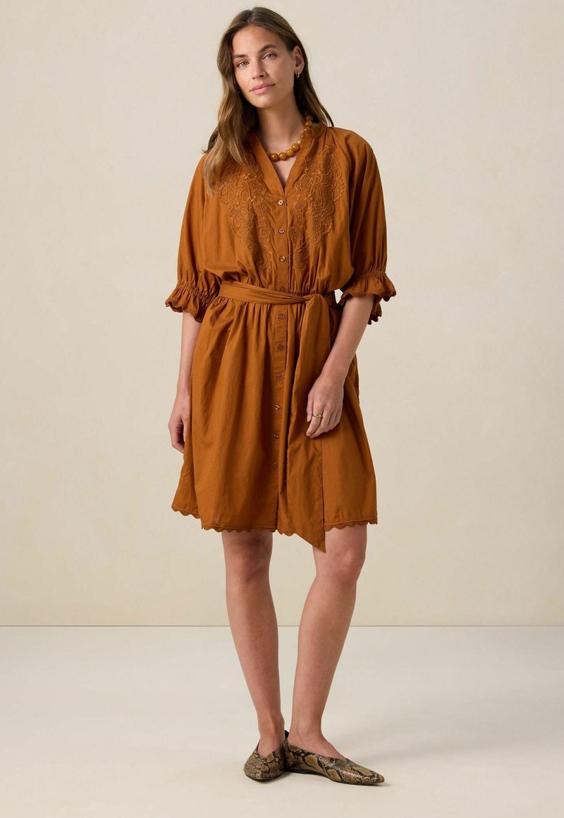 Rostbruine jurk met korte mouwen, knoopsluiting aan de voorkant, geborduurde details, een ceintuurlijn en een geschulpte zoom; gladde textuur.