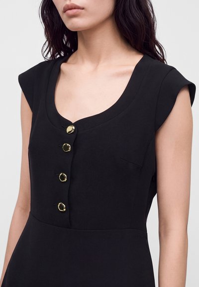 Femme portant une robe noire sans manches avec un décolleté rond et quatre boutons décoratifs dorés sur le devant.