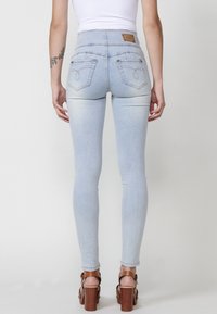 Vaqueros ajustados de color azul claro en denim elástico, con bolsillos traseros con costuras decorativas y un diseño de cintura alta, acompañados de tacones de plataforma marrones.