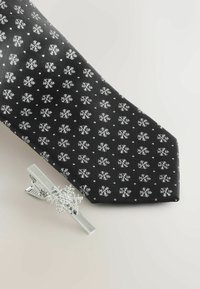 Fekete nyakkendő fehér hópehely mintával és apró ezüst nyakkendőcsipke hópehely formával. Sima anyag textura, kecses fazon.