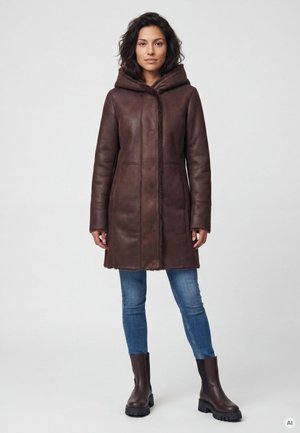 Frau trägt einen braunen Lederkapuzenmantel, blaue Jeans und dunkelbraune Stiefel, steht vor einem schlichten grauen Hintergrund.