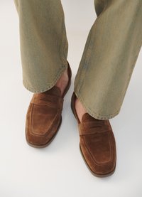 Brūni zamšādas loafers ar gludu apdari, kvadrātveida purngalu un dekoratīvu šuvumu, apvienoti ar gaiši zaļām, viegli izplestām biksēm.