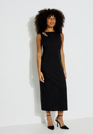 Femme aux cheveux bouclés portant une robe midi noire sans manches avec des perles décoratives près du décolleté et des chaussures à talons hauts noires.