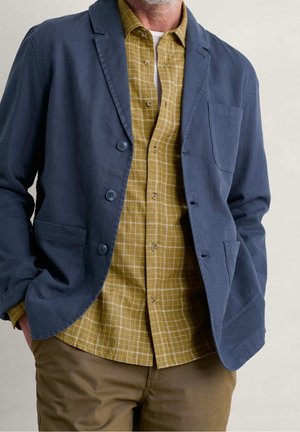 Homme portant un blazer bleu, une chemise à carreaux jaune moutarde et un pantalon marron, les mains dans les poches, devant un fond uni.