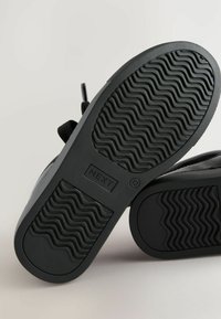 Schwarze Gummisneaker mit einer strukturierten Sohle, die wellenförmige Muster und Branding aufweist. Die Schnürsenkel sind oben sichtbar und haben ein schlichtes Design.