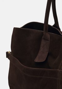 Sac fourre-tout en daim brun avec un intérieur spacieux, une grande ouverture et une poche extérieure. Doté d'une sangle cousue et d'accents de quincaillerie minimalistes.