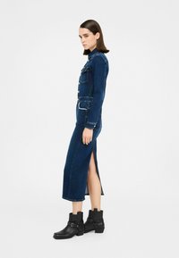 Abito di jeans con maniche lunghe, colore blu scuro, tasche frontali e spacco laterale. Abbinato a stivaletti neri alla caviglia con cinturini.