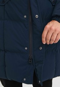 Tamno plavi puffer jakna s zatvaranjem na zip, kopčama na dugmad i kviltanim teksturom. Ima stojeći ovratnik i blago prilagođen kroj.