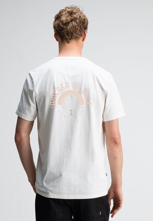 Blonder Mann mit kurzen, lockigen Haaren, der ein weißes T-Shirt mit kurzen Ärmeln trägt, auf dessen Rücken sich ein hellbeiger, kreisförmiger Text und eine Grafik befinden.