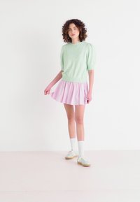 Mintgrön kortärmad tröja i kombination med en rosa plisserad kjol. Modellen bär vita knästrumpor och ljusgröna sneakers. Slät textur.