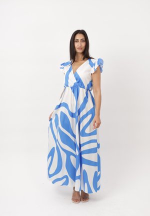 Mujer con un vestido maxi blanco sin mangas con patrones abstractos azules, de pie frente a un fondo blanco liso.