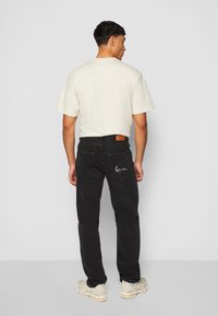 Black jeans with a subtle white logo on the back pocket, paired with a cream-colored short-sleeve t-shirt. Casual, relaxed fit.