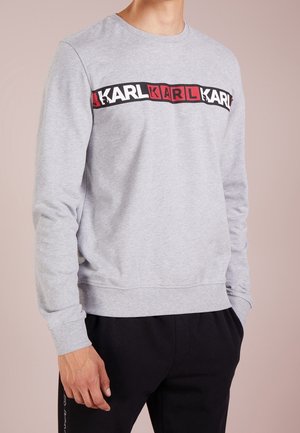 Homme portant un sweat-shirt gris clair à manches longues avec le logo « KARL » répété en blocs blancs et rouges sur la poitrine, associé à un pantalon noir.
