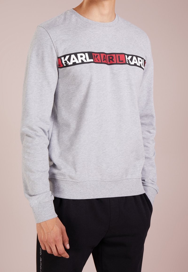 Homme portant un sweat-shirt gris clair à manches longues avec le logo « KARL » répété en blocs blancs et rouges sur la poitrine, associé à un pantalon noir.