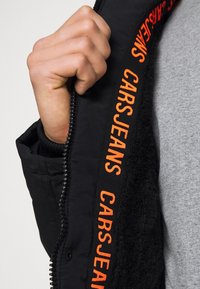 Svart jacka med mjukt innerfoder. Har kraftig orange "CARS JEANS" märkning vid dragkedjans kant. Dragkedjan är silverfärgad och funktionell.