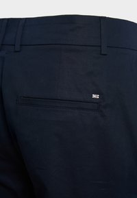 Marineblauwe broeken met een platte voorkant, een achterzak met bies en een klein logoaccent nabij de zak. Het materiaal is soepel en gestructureerd.