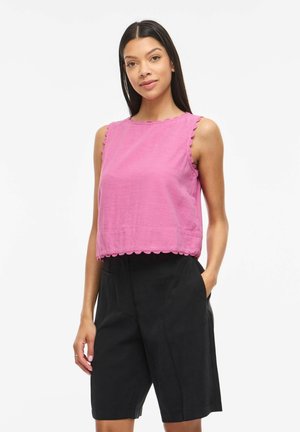 Femme portant un haut rose sans manches à bords festonnés et un short noir jusqu'aux genoux, debout avec une main dans la poche sur fond blanc.