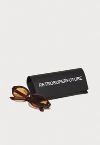 Ochelari de soare maro cu model tortoiseshell, cu rame rotunde, plasati langa un capac de protectie negru marcat cu "RETROSUPERFUTURE" in alb.