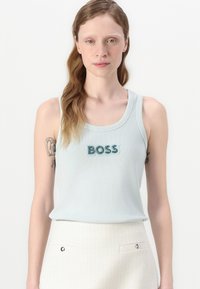 Svetlo modra rebrasta majica na naramnice s teksturiranim logotipom "BOSS" v temnejši odtenek. V kombinaciji z belo teksturirano krilom.