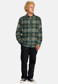 Camicia flanella a righe in tonalità blu, verde e marrone, con colletto boton-down e maniche lunghe. Indossata con pantaloni scuri e sneakers.