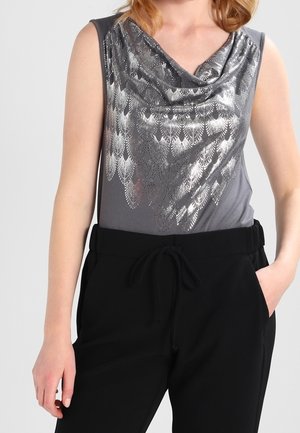 Szary top bez rękawów z metalicznym, srebrnym wzorem przypominającym pióra, w połączeniu z czarnymi spodniami z sznurkiem, jedna ręka w kieszeni.