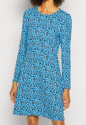 Frau trägt ein blaues Kleid mit langen Ärmeln und einem schwarzen und orangefarbenen Leopardenmuster, dargestellt von Hals bis über die Knie.