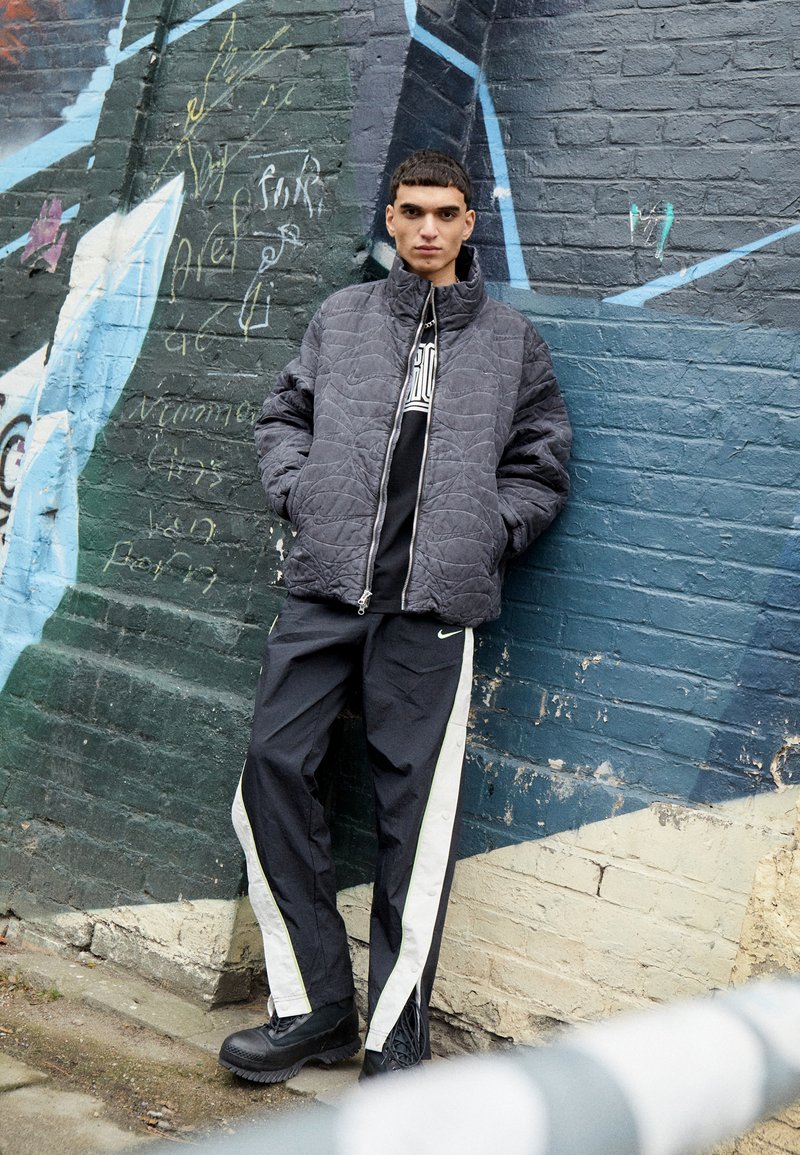 Giacca grigia trapuntata con colletto alto, felpa nera sotto, pantaloni neri con strisce bianche ai lati e stivali neri chunky, davanti a un muro di graffiti.