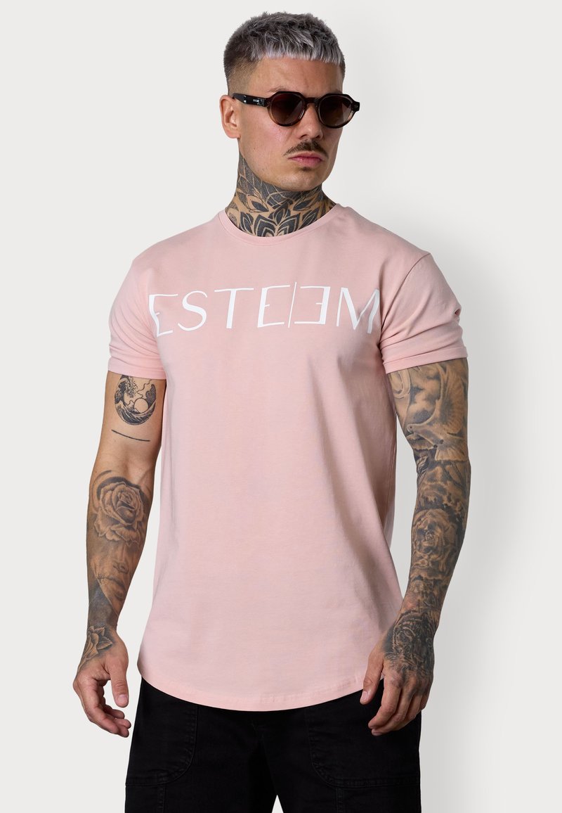 Hellrosa T-Shirt mit kurzen Ärmeln und dem weißen Aufdruck "ESTEEM". Verfügt über einen Rundhalsausschnitt und einen leicht geschwungenen Saum. Schwarze Hose und Sonnenbrille.