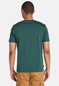 Muž stojící zády, oblečený v tmavě zeleném tričku s krátkým rukávem a béžových cargo šortkách na jednobarevném bílém pozadí.