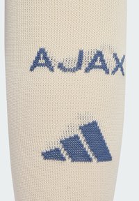 Sportske čarape boje krem sa plavim pletenim natpisom "AJAX" i plavim grafičkim motivom s tri pruge na dnu, izrađene od prozračnog materijala.