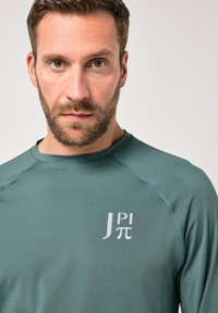 Teal urheilu pitkähihaisia paitoja, joissa on ribbikuvio, kevyesti harmaan värinen logo rinnassa ja kontrastivärjäystä olkapäissä.