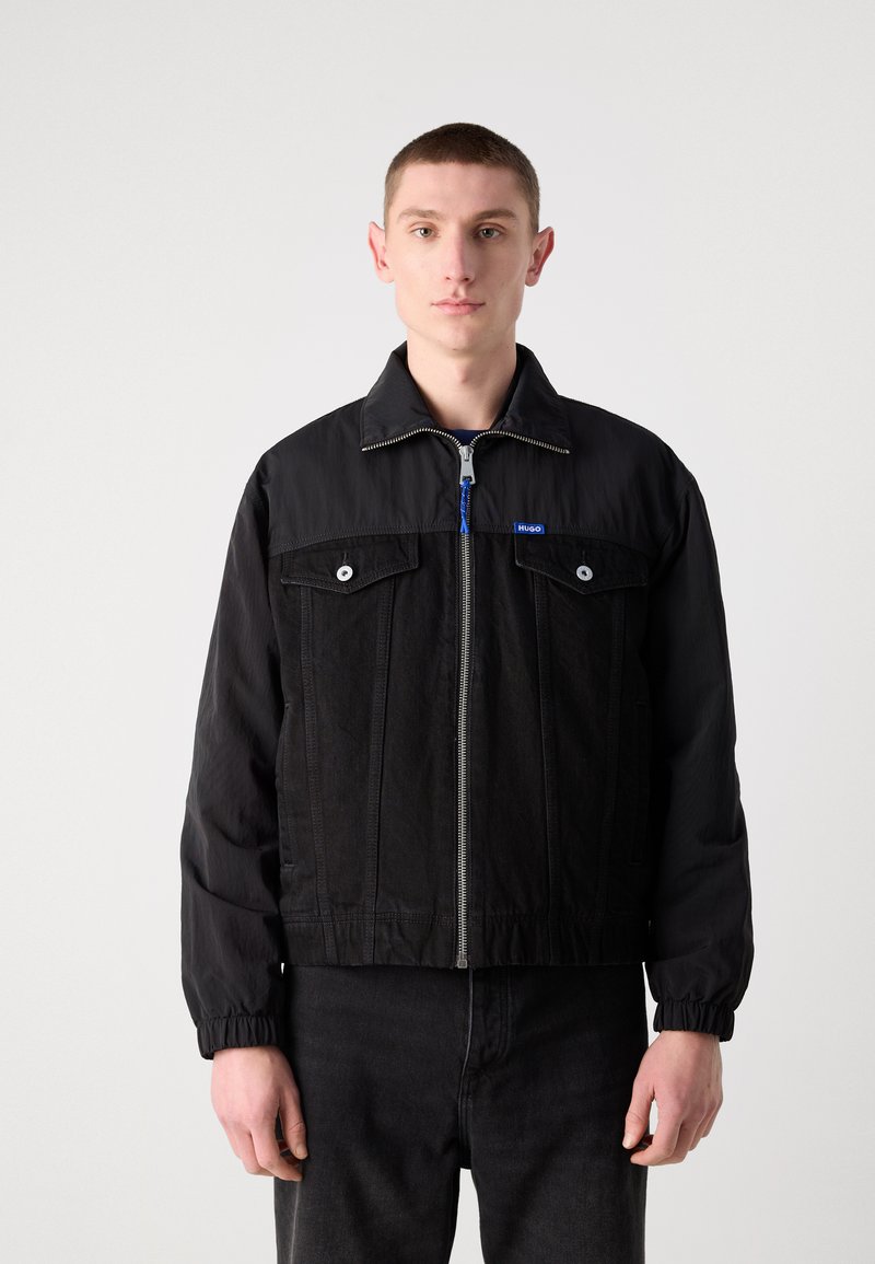 Czarna kurtka z kombinacją gładkiego materiału i teksturowanej dżinsówki, z zapięciem na zamek z przodu, dwoma kieszeniami na piersi i elastycznymi mankietami.