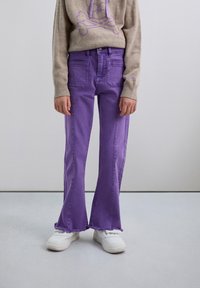 Jeans morados acampanados con un dobladillo sin terminar, hechos de tela vaquera. Combinados con zapatillas blancas y un suéter beige con un diseño de calavera.