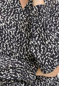 Schwarz-weißer gemusterter Stoff mit einer zerknitterten Textur. Verfügt über einen V-Ausschnitt und puffige Ärmel, die an den Manschetten gerafft sind.