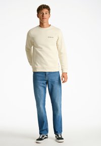 Sudadera color crema con textura acanalada y detalle de logo, combinada con jeans de mezclilla azules y zapatillas negras con acentos blancos.