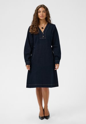 Femme aux cheveux bouclés portant une robe en denim sombre à manches longues et des talons pointus noirs, debout devant un fond uni.