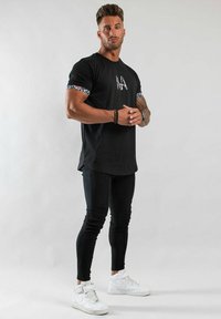 Svart t-shirt med blommönster på ärmarna, kombinerad med svarta leggings och vita sneakers. Slät textur, avslappnad passform, logotyp på bröstet.