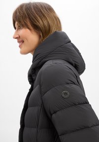 Schwarze Pufferjacke mit hohem Kragen und einem runden Logo-Patch auf der linken Schulter. Das Design verfügt über gesteppten Bereiche und eine glatte Textur.