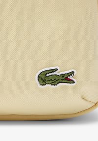 Gelber, strukturierter Stoffbeutel mit einem gestickten grünen Krokodil-Logo und roten Akzenten. Kompakte Form mit abgerundeten Kanten.