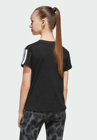 T-shirt en coton noir à manches courtes avec des rayures blanches triples sur l'épaule gauche, associé à des leggings à motifs noir et gris.