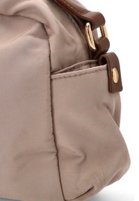 Esquina de un bolso de tela beige con correa de cuero marrón y hebilla de metal dorado, mostrando detalles de costura y un bolsillo lateral.
