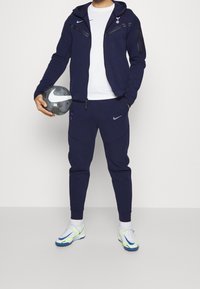 Navy tracksuit with zippered pockets, featuring a white T-shirt beneath. Holding a black and white Nike soccer ball. White and neon shoes.