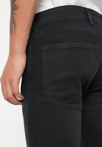 Jeans in denim nero con una tasca posteriore caratterizzata da un motivo di cucitura a zigzag, vita alta e texture liscia. Mano appoggiata sul lato.