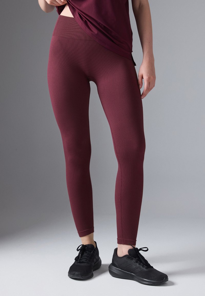 Colanți cu un design texturat de culoare vișinie, cu talie înaltă, care se potrivesc foarte bine. Asortați cu încălțăminte sport negre pe un fundal gri deschis.