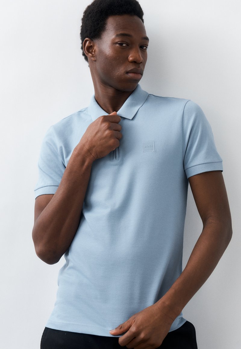 Νεαρός άνδρας που φοράει ανοιχτό γαλάζιο πόλο πουκάμισο, ρυθμίζοντας το γιακά με το ένα χέρι, στέκεται μπροστά σε απλό λευκό φόντο.