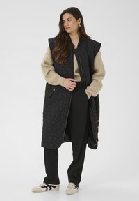 Long gilet matelassé noir avec fermeture éclair à l'avant, design oversize, poches latérales, assorti à un pull beige et un pantalon noir, avec des baskets crème.
