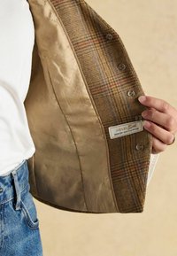 Vnútro tweedového saka s béžovou saténovou podšívkou, ozdobené jemným vzorom a štítkom s nápisom "JOULES Tweed."